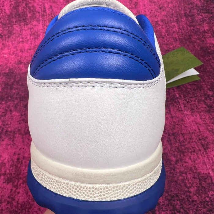 Sneaker bianca con dettagli blu e tag verde su sfondo rosa vibrante.