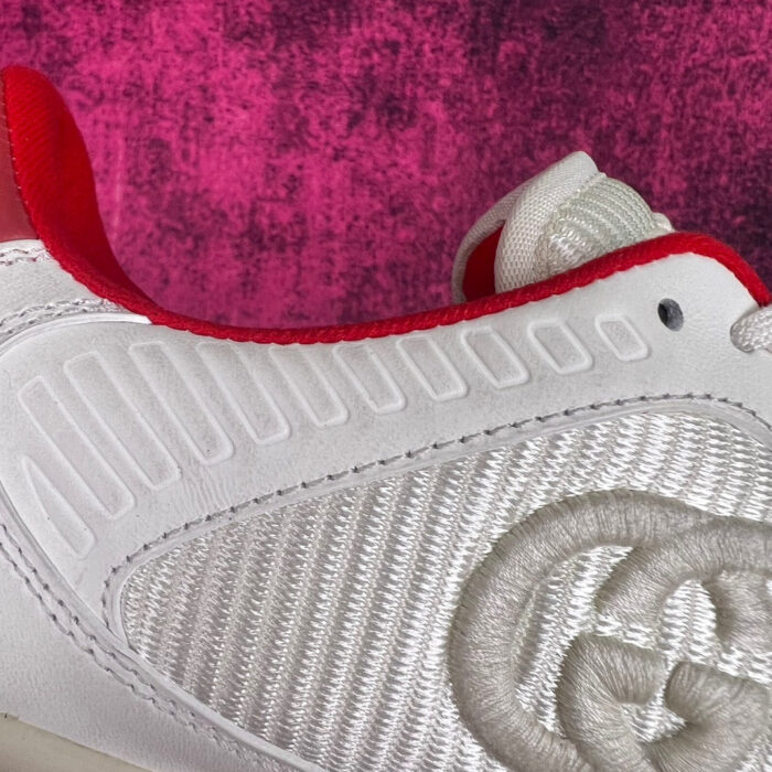 Sneaker moderna bianca con dettagli ricamati e accenti rossi, su sfondo viola textured.