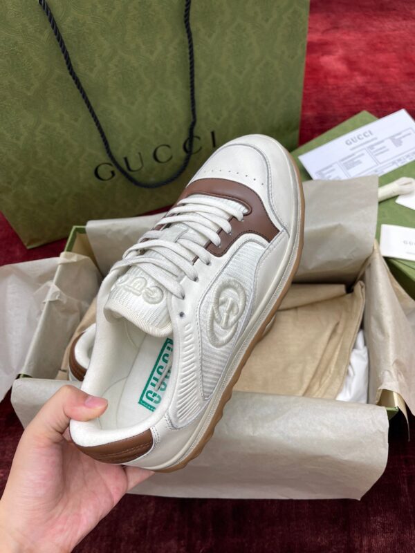 Sneaker bianca di lusso con dettagli marroni, in confezione elegante e sfondo rosso raffinato.