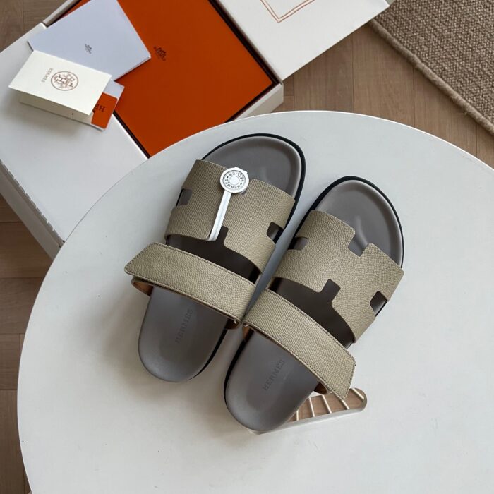 Sandali di lusso con logo H, stile moderno, su tavolo bianco, packaging elegante e design minimalista.