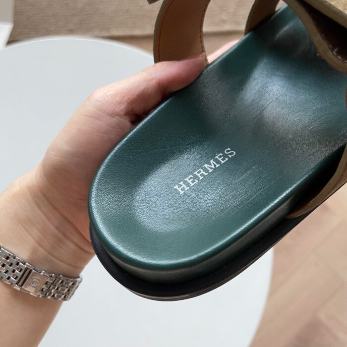 Sandalo Hermes di lusso in pelle verde con logo, mano con orologio elegante.