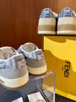 Sneakers di lusso Fendi con dettagli in tessuto e suede su supporto trasparente, scatola gialla elegante.