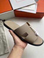 Sandali beige minimalisti con dettaglio H, su suola nera, in cornice di confezione luxury.