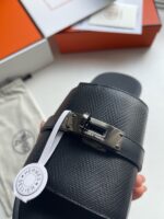 Sandalo slip-on in pelle nera con fibbia metallica, brand di lusso su sfondo bianco e scatola arancione.