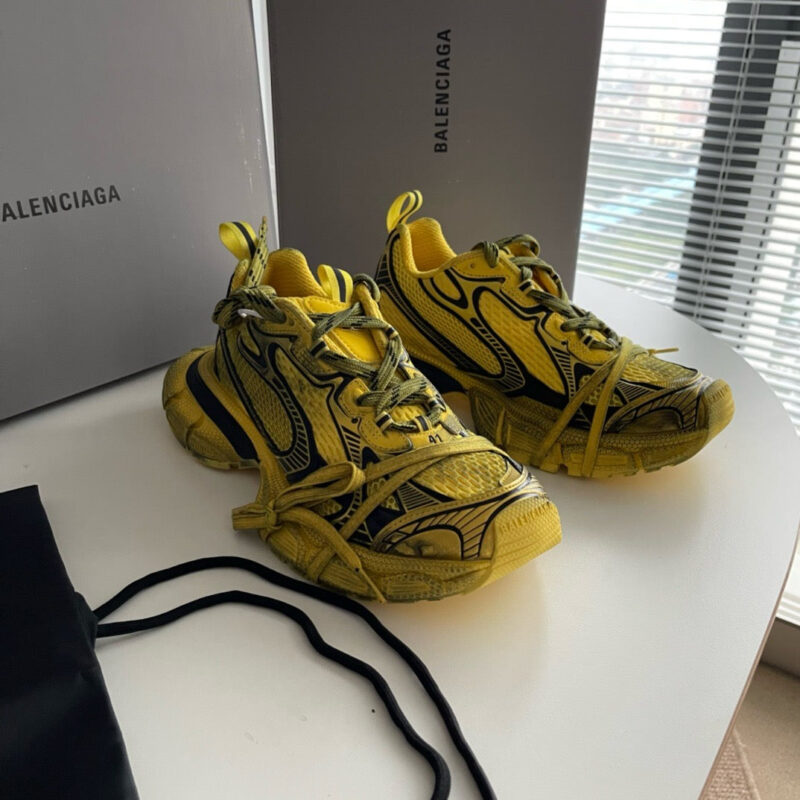 Sneakers gialle Balenciaga in esposizione elegante su tavolo bianco con scatole e borsa nera.