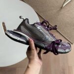 Scarpa sportiva grigia con dettagli viola, tenuta in mano su pavimento in legno naturale.