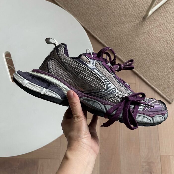 Scarpa sportiva grigia con dettagli viola, tenuta in mano su pavimento in legno naturale.