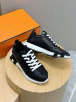 Sneakers di lusso in pelle nera con lacci bianchi su supporto in legno e scatola arancione.