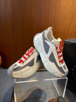 Sneakers moderne con dettagli grigi e lacci rossi, esposte su pedana di vetro con sfondo in legno.