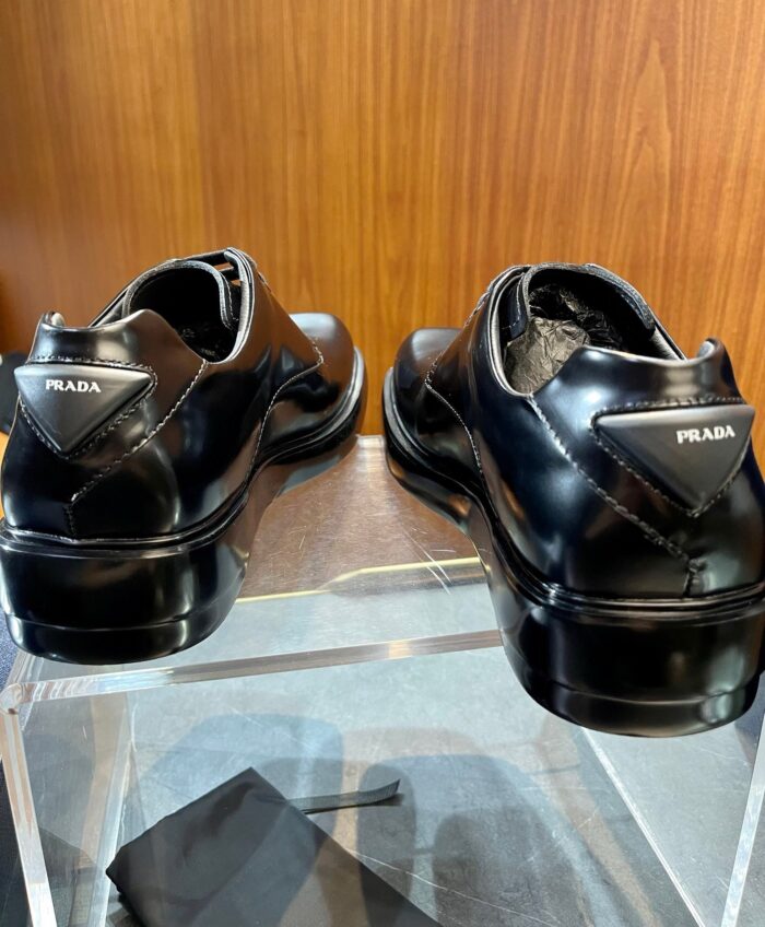 Scarpe da uomo eleganti in pelle nera con logo triangolare su sfondo in legno luccicante.