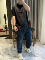 Persona con polo nero, jeans blu e sneakers in ambientazione casalinga luminosa.