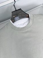 Felpa grigia casual con scollo rotondo e layering, su appendiabiti trasparente minimalista.