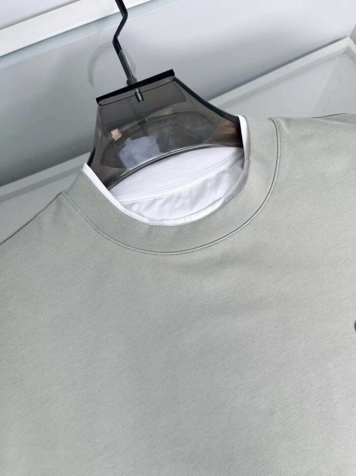Felpa grigia casual con scollo rotondo e layering, su appendiabiti trasparente minimalista.