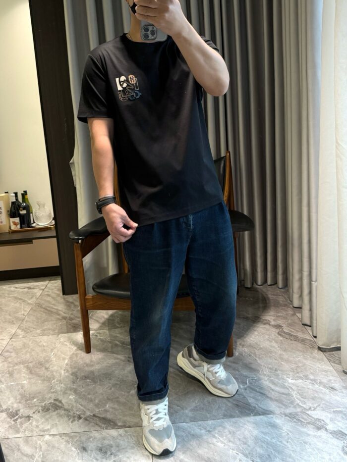 Abbigliamento streetwear casual con maglietta nera, jeans e sneakers colorati in ambiente domestico.