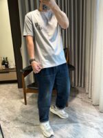 Uomo in casa con jeans, t-shirt grigia e sneakers bianchi, selfie casual e rilassato.