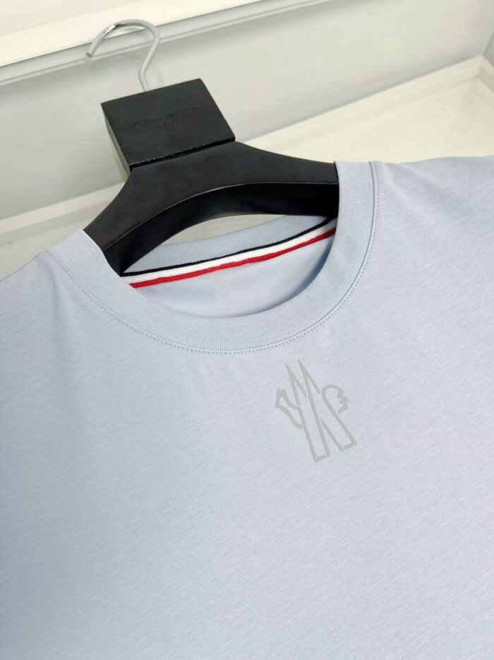 Maglietta blu chiara, logo minimale, dettaglio interno a strisce rosso, bianco e blu.