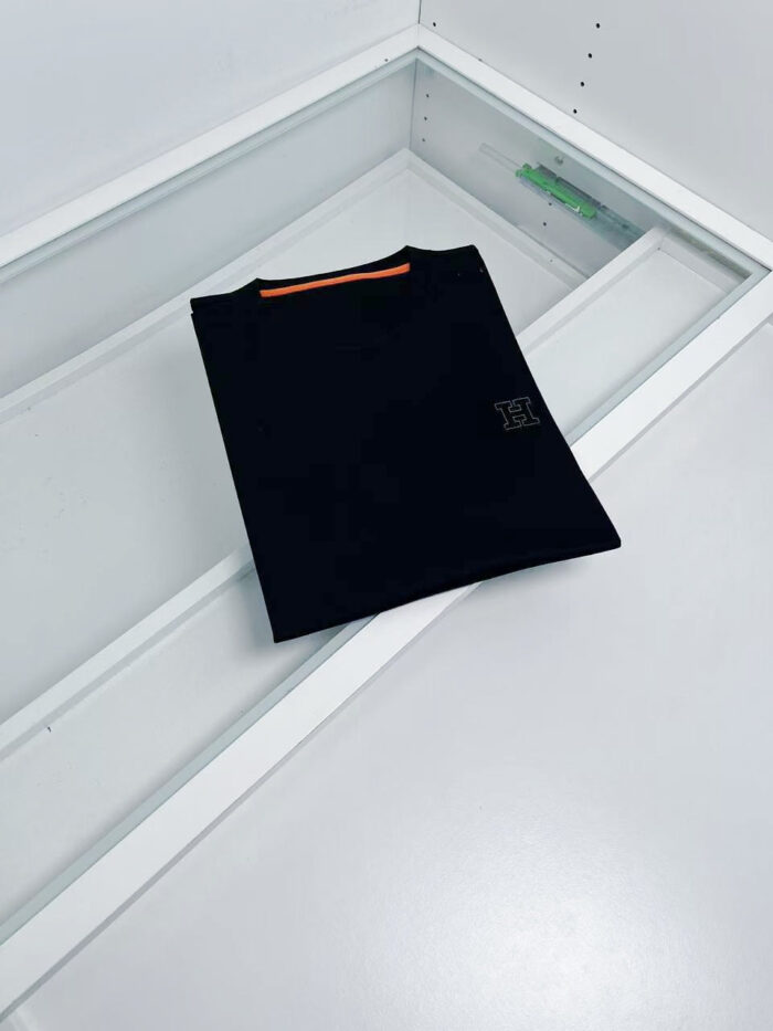 Maglietta nera minimalista con striscia arancione e logo discreto, display elegante su sfondo bianco.