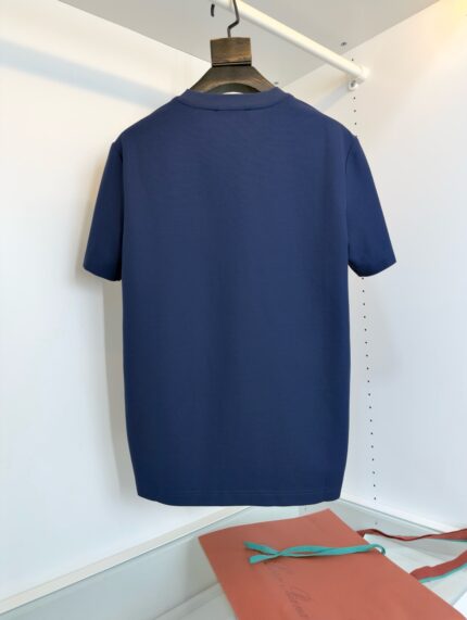 Maglietta navy blue minimalista con dettaglio arancione e accessorio blu, su appendiabiti di legno.