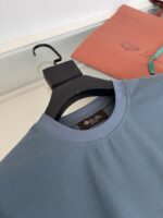 Maglietta azzurra semplice su appendiabiti nero, con dettaglio elegante e sfondo sfocato.