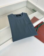 Maglietta blu semplice e comoda su superficie lucida, con sacchetto regalo colorato sullo sfondo.