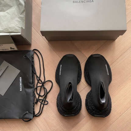 Scarpe Balenciaga nere con packaging di lusso su pavimento in legno chiaro.
