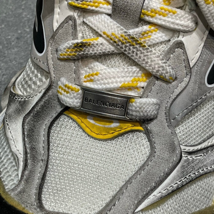 Sneaker Balenciaga grigia, bianca e gialla, con logo in metallo e suola sportiva.