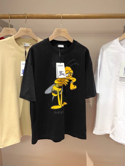 Maglietta nera con grafica ape giocosa, esposta tra magliette beige e bianche su supporto in legno.
