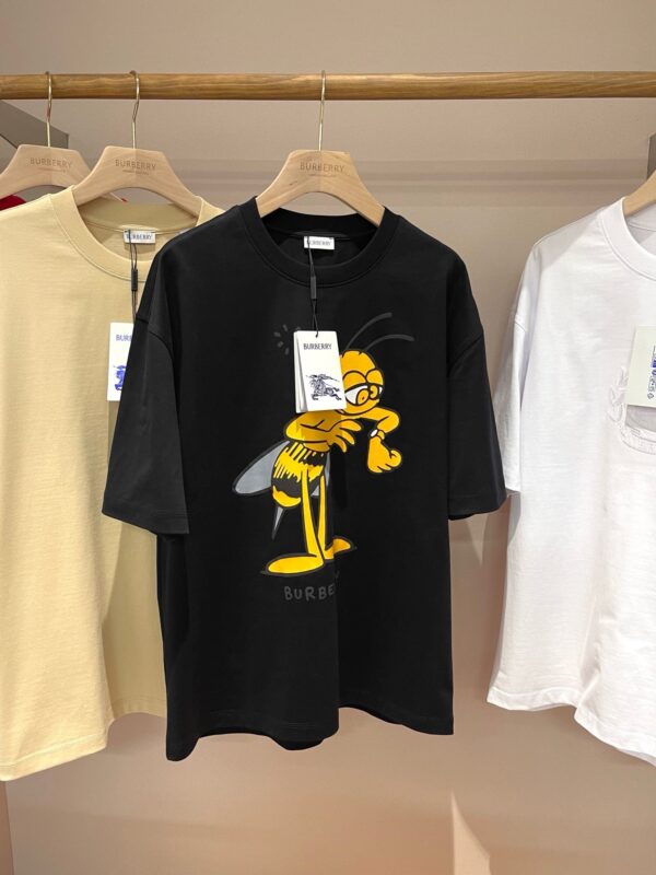 Maglietta nera con grafica ape giocosa, esposta tra magliette beige e bianche su supporto in legno.