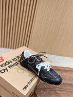 Sneaker New Balance nera con lacci bianchi su scatola e pavimento in legno naturale.