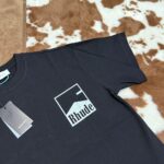 Maglietta nera Rhude con logo minimalista su sfondo in pelle animale, stile streetwear moderno.