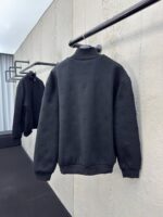 Maglione nero minimalista appeso in setting moderno con parete di cemento e luce soffusa.