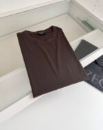 Maglietta marrone semplice e elegante, piegata su superfice bianca minimalista, stile casual comfort.