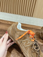 Sneaker New Balance in suede marrone con lacci arancioni, logo N e dettagli artigianali.