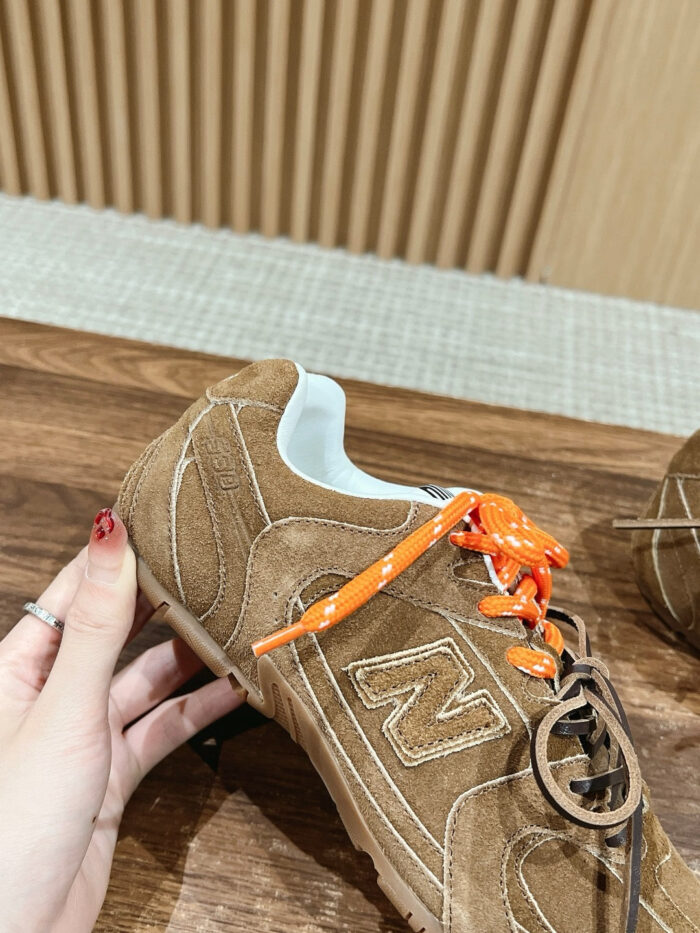 Sneaker New Balance in suede marrone con lacci arancioni, logo N e dettagli artigianali.