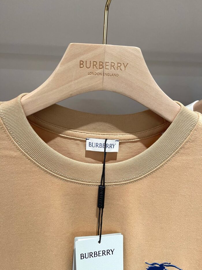 Camicia beige Burberry con ricamo logo, design elegante e dettagli di lusso su gancio di legno.
