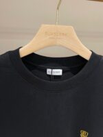 Maglietta nera Burberry con logo giallo, elegante e versatile, su appendiabiti di legno lucido.