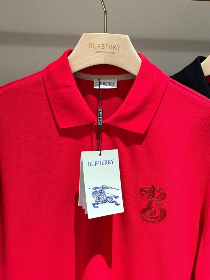 Polo rossa Burberry con logo ricamato e etichetta elegante su appendiabiti in legno.