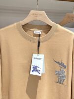 T-shirt Burberry beige con stampa di cavaliere medievale blu, casual ed elegante, su gruccia di legno.