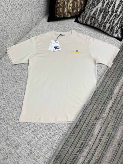 T-Shirt beige casual con ricamo giallo, su divano con cuscini decorativi, stile minimal e confortevole.