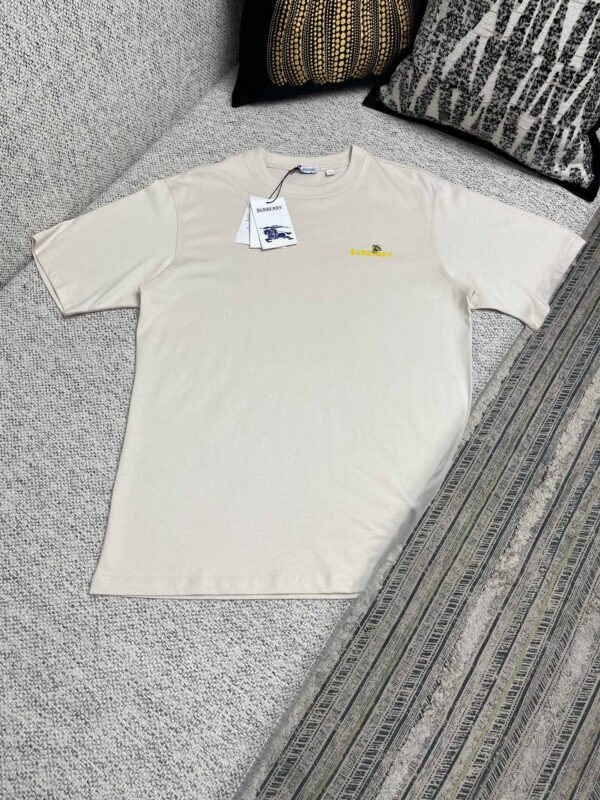 T-Shirt beige casual con ricamo giallo, su divano con cuscini decorativi, stile minimal e confortevole.