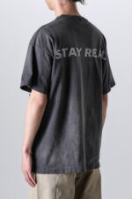 Maglietta nera oversize con slogan STAY REAL e pantaloni beige, stile casual e vintage.