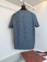 Camicia blu casual su appendiabiti in ambiente minimalista con sacchetto ruggine e tracolle verdi.