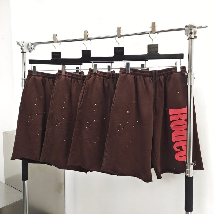 Pantaloncini casual marroni con ritagli e logo rosso, stile streetwear moderno e giovanile.
