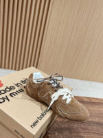 Scarpa in camoscio marrone con lacci bianchi e scatola New Balance su sfondo di legno.