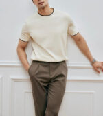 Uomo elegante casual con camicia crema e pantaloni marroni in ambiente minimalista.
