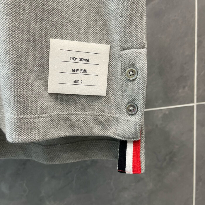 Maglietta polo grigia con etichetta moderna, bottoni metallici e dettaglio a strisce colorate.