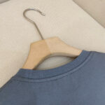 Camicia grigia casual su appendiabiti in legno con sfondo neutro e stile minimalista.