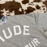 Maglietta grigia con logo NUDE su sfondo a pelo di mucca, stile moderno e casual.