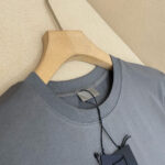 T-shirt grigia casual, cotone leggero, scollo rotondo, su appendiabiti in legno chiaro.
