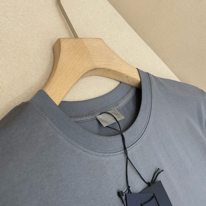 T-shirt grigia casual, cotone leggero, scollo rotondo, su appendiabiti in legno chiaro.
